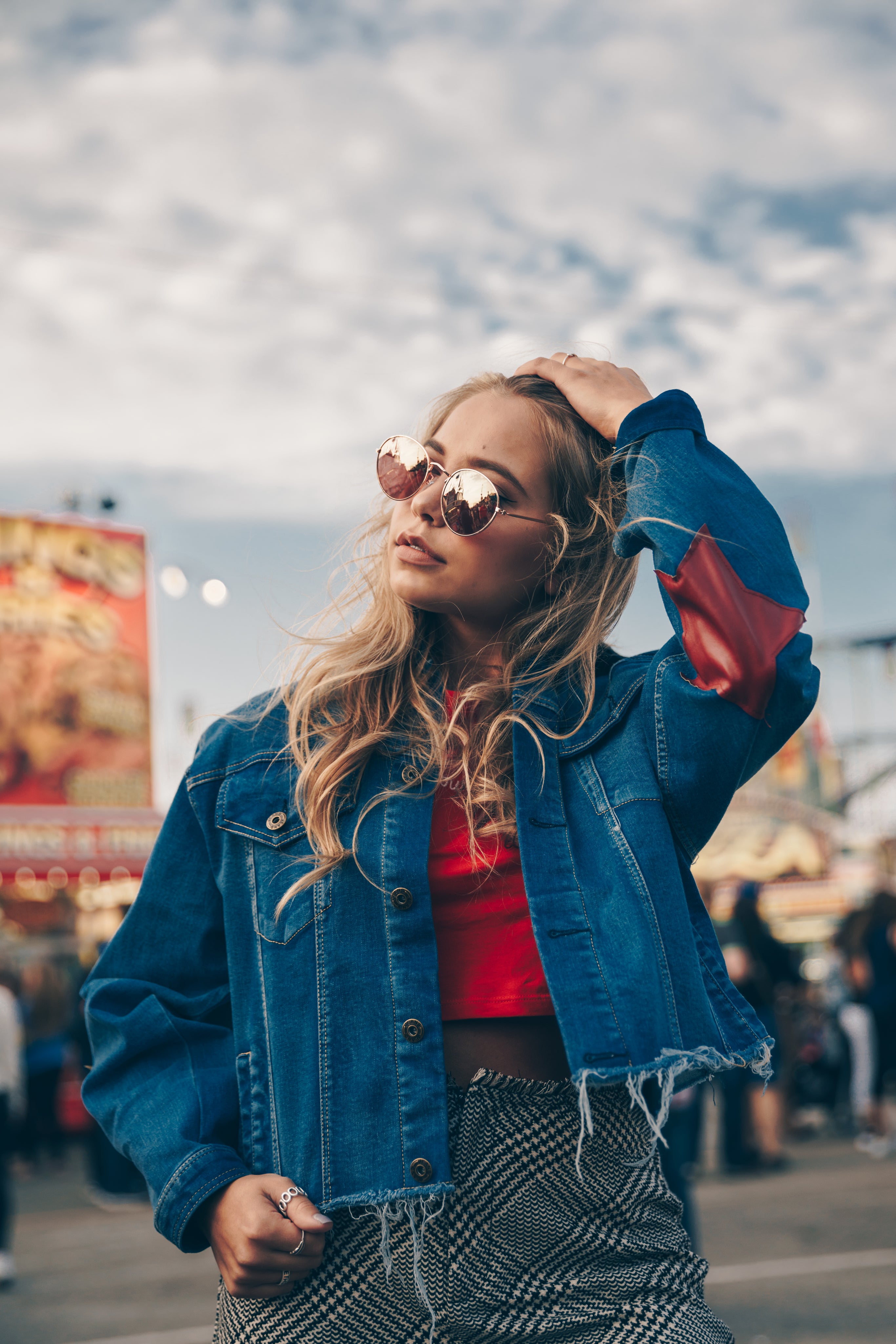 White woman wearing black jeans, red shirt, and frayed jean jacket with red star on elbow. She has long blonde hair and wearing mirrored sunglasses.