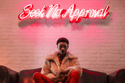 Black woman wearing cream fur coat with orange pants sitting on brown low back leather couch. She is in front of a white brick wall with a hot pink neon sign that says seek no approval